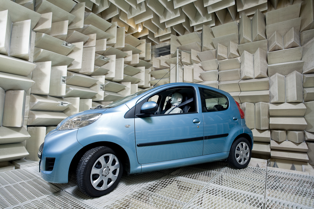 Blue passenger car with a test dummy in the driver's seat, positioned inside an anechoic chamber with acoustic wedges on the walls and ceiling and a metal mesh floor.